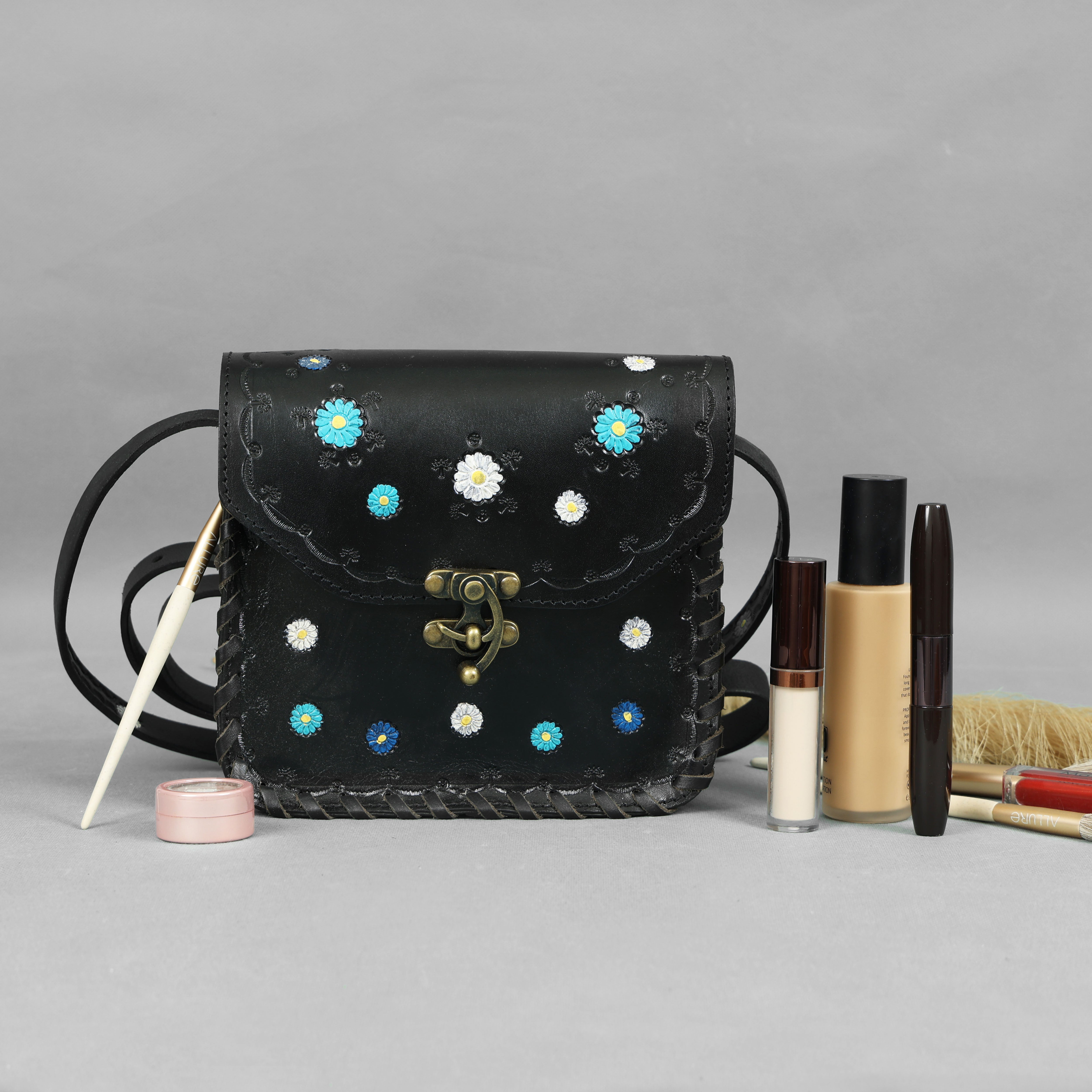 Black handbag with floral patterns next to makeup items on a gray background
