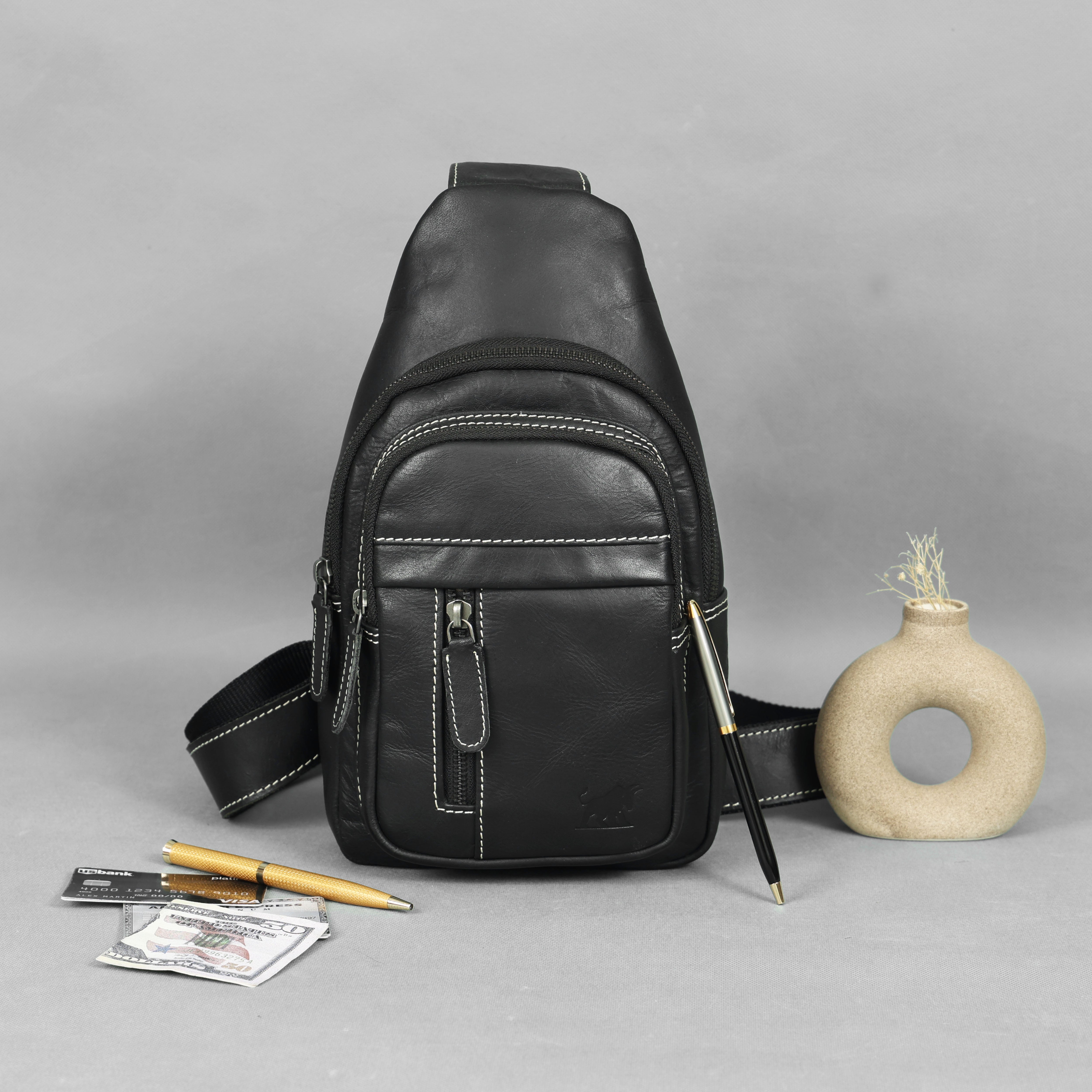 Black leather sling bag on a gray background with a pen, money, and small vase.