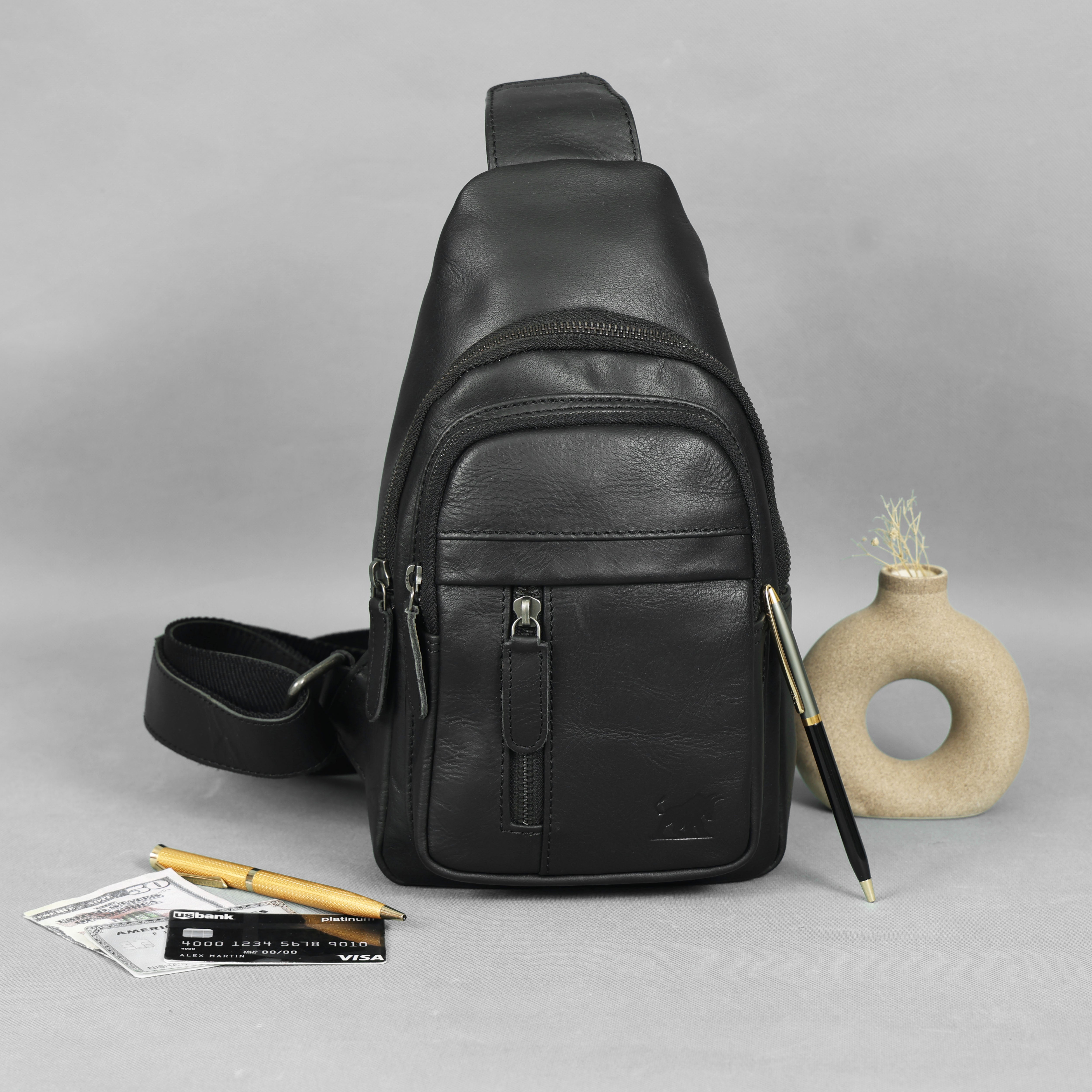 Black leather sling bag on a gray background with a pen and small plant.