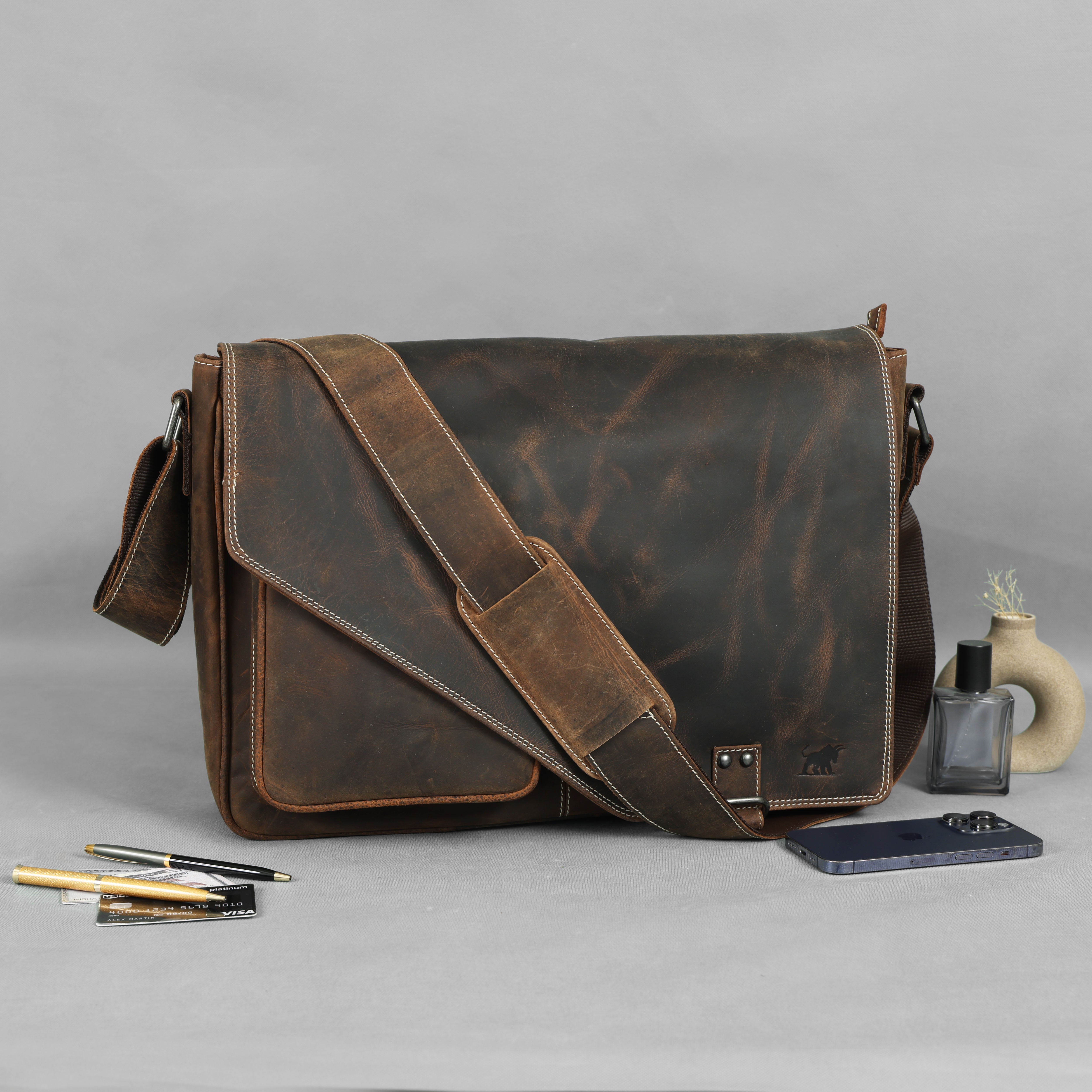 Brown leather messenger bag on a gray surface with stationery items.
