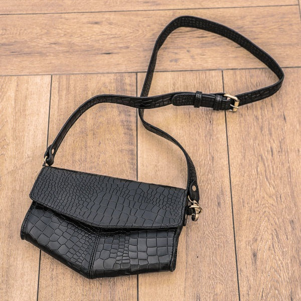 A black crocodile pattern leather sling bag with an adjustable strap placed on a wooden floor.