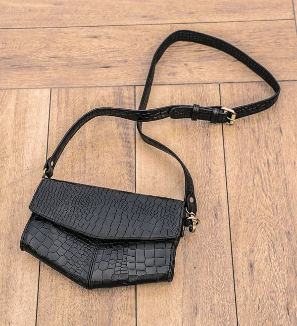 A black crocodile pattern leather sling bag with an adjustable strap placed on a wooden floor.