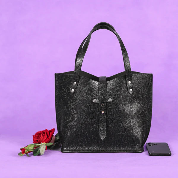 A black leather tote bag with embossed patterns, displayed against a purple background with a red rose and a smartphone beside it.