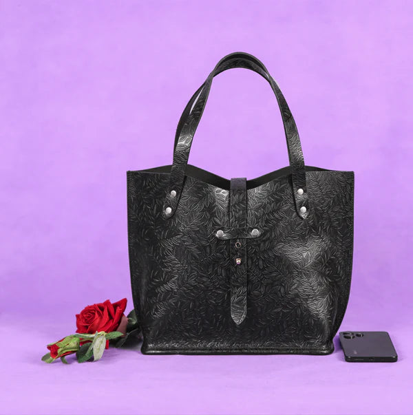 A black leather tote bag with embossed patterns, displayed against a purple background with a red rose and a smartphone beside it.