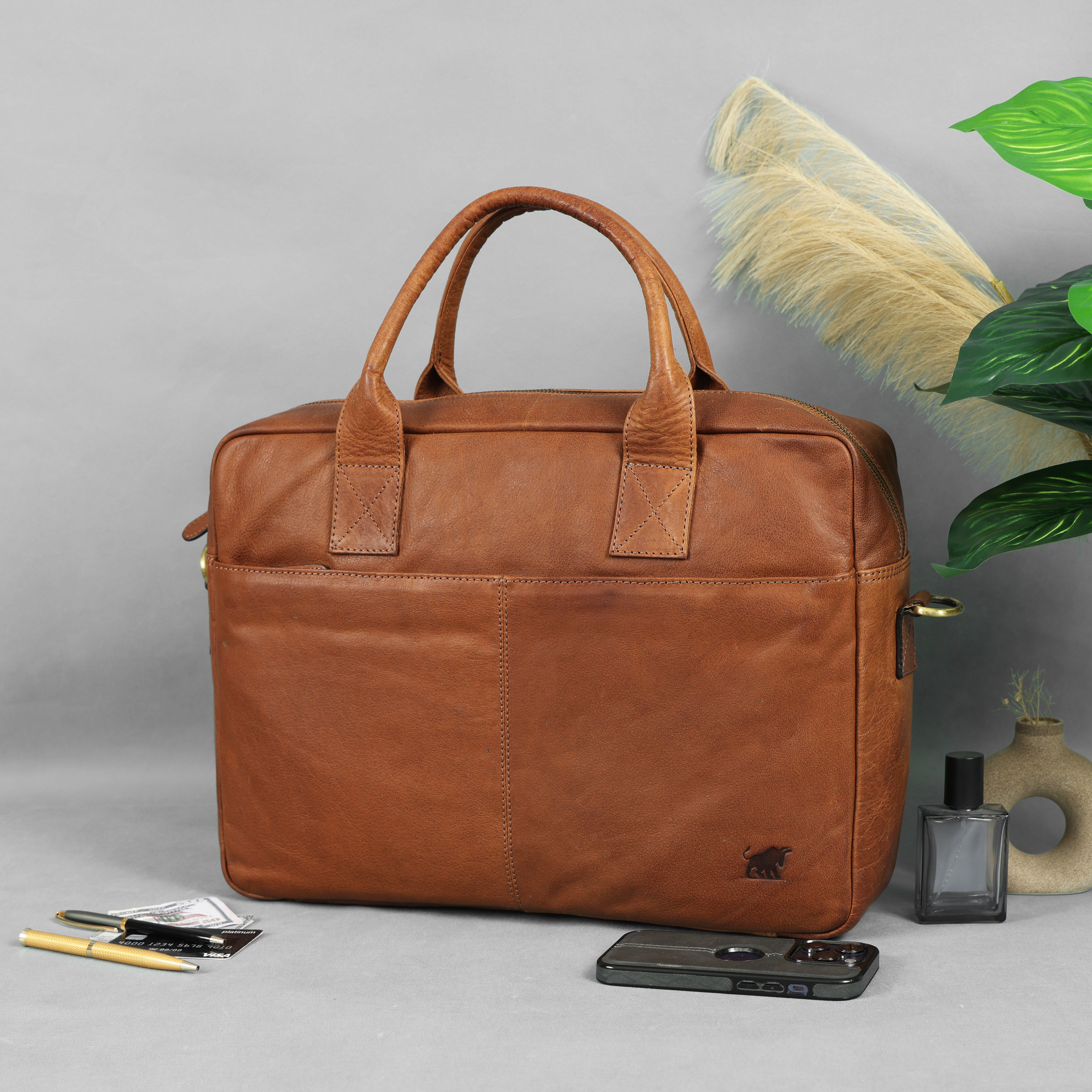 Washed brown leather laptop bag with a removable shoulder strap, handles, and two front pockets, displayed on a grey surface with a smartphone, pens, and decorative items.