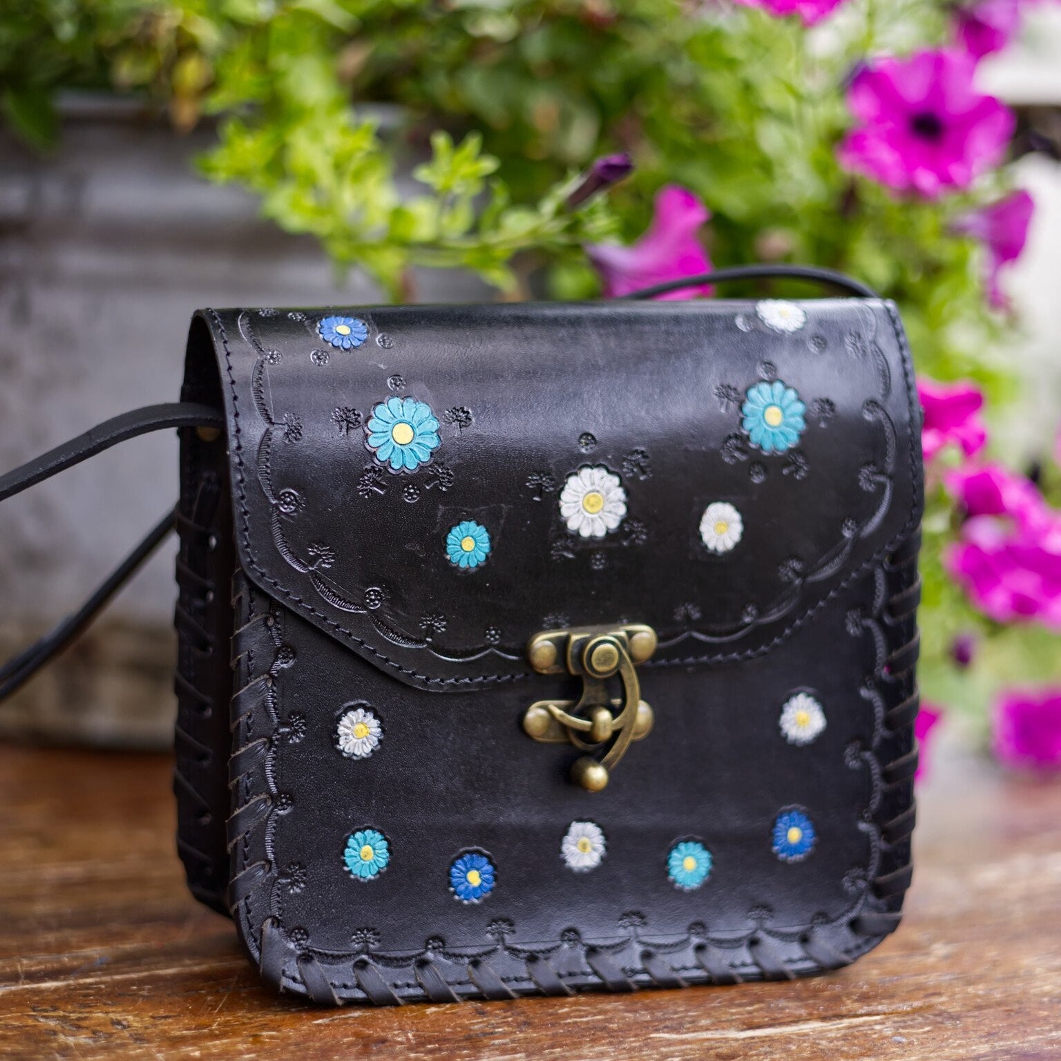 Black leather bag with floral patterns on a wooden surface with flowers in the background
