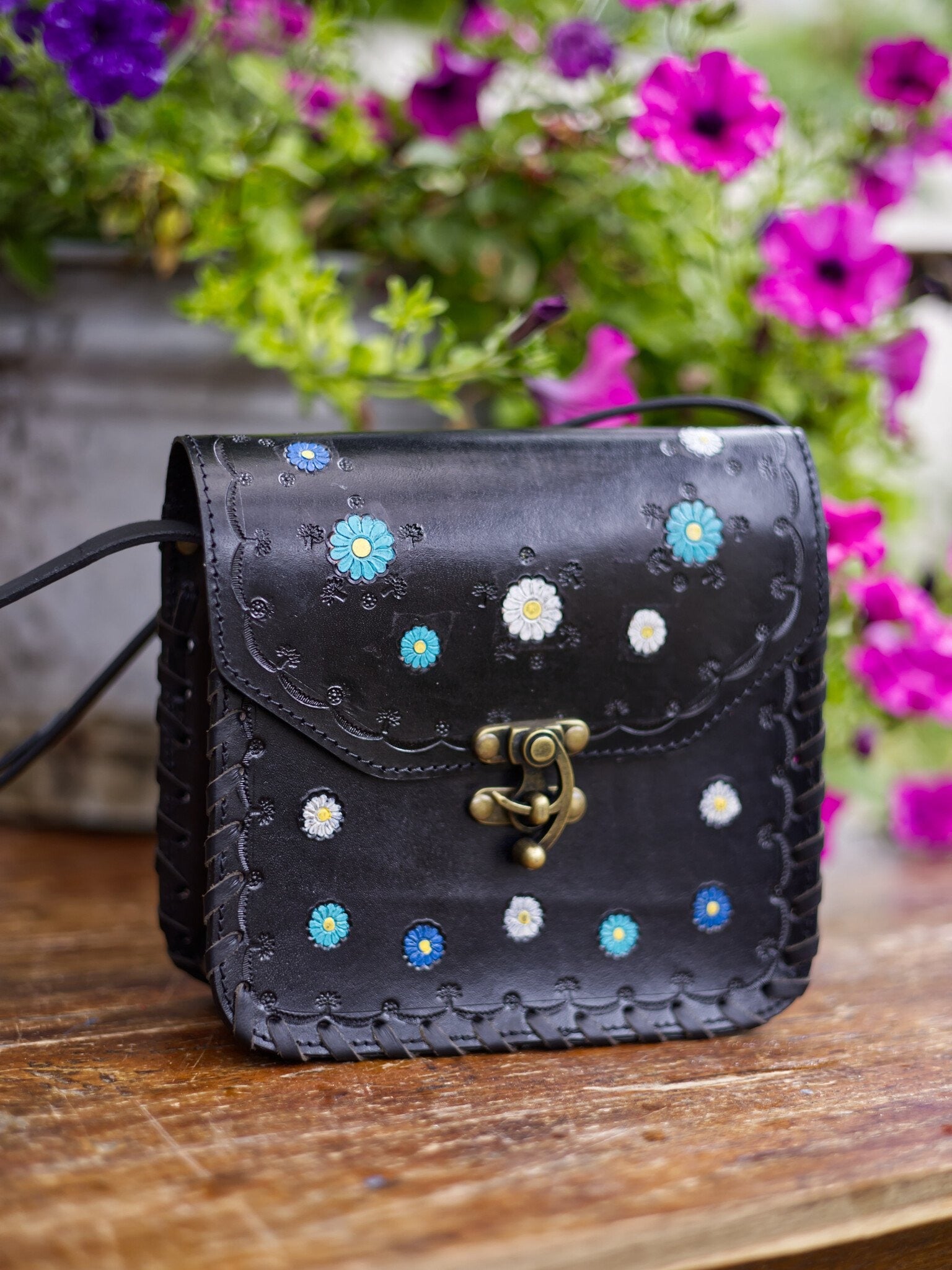 Black leather bag with floral patterns on a wooden surface with flowers in the background