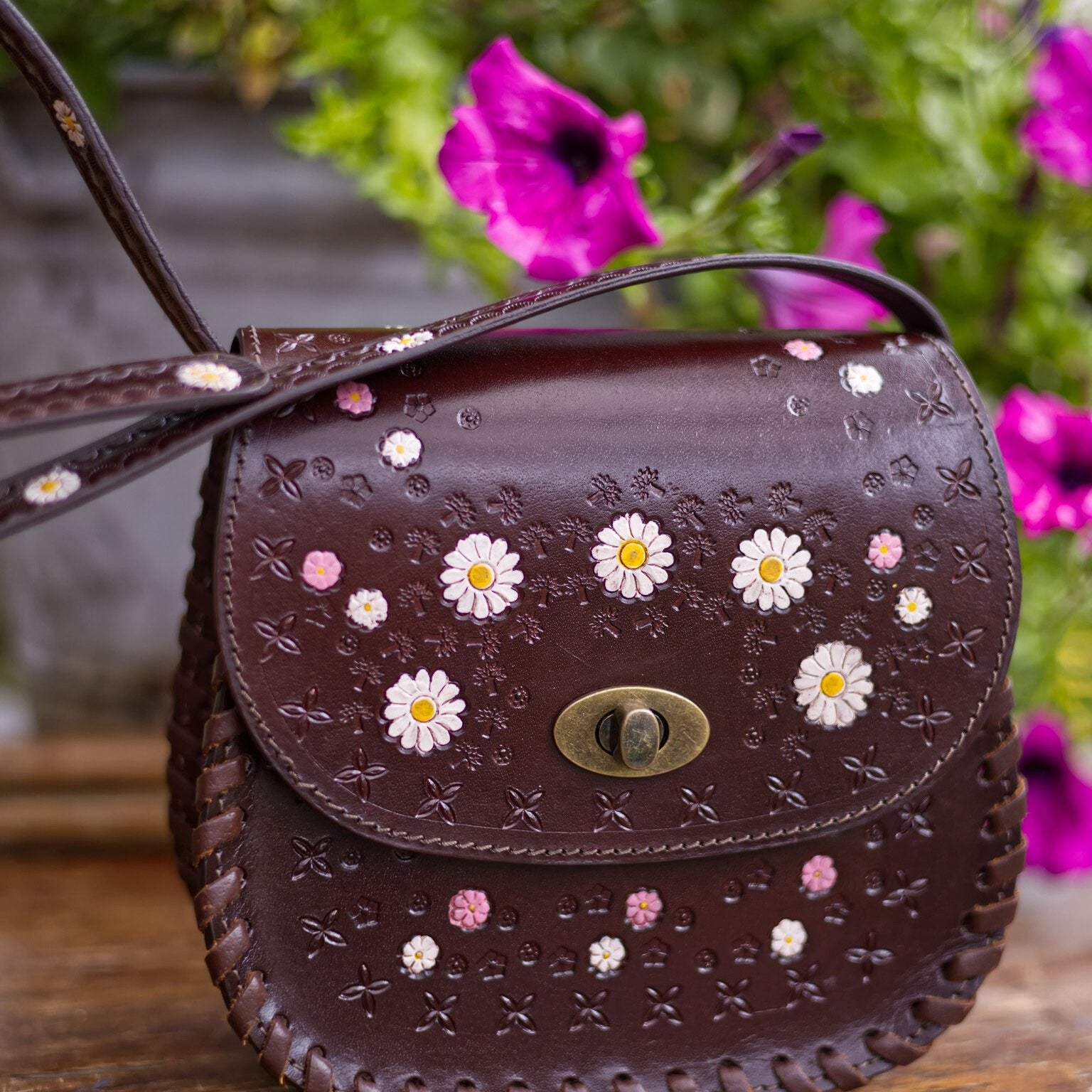 Brown leather handbag with floral patterns on a wooden surface with flowers in the background