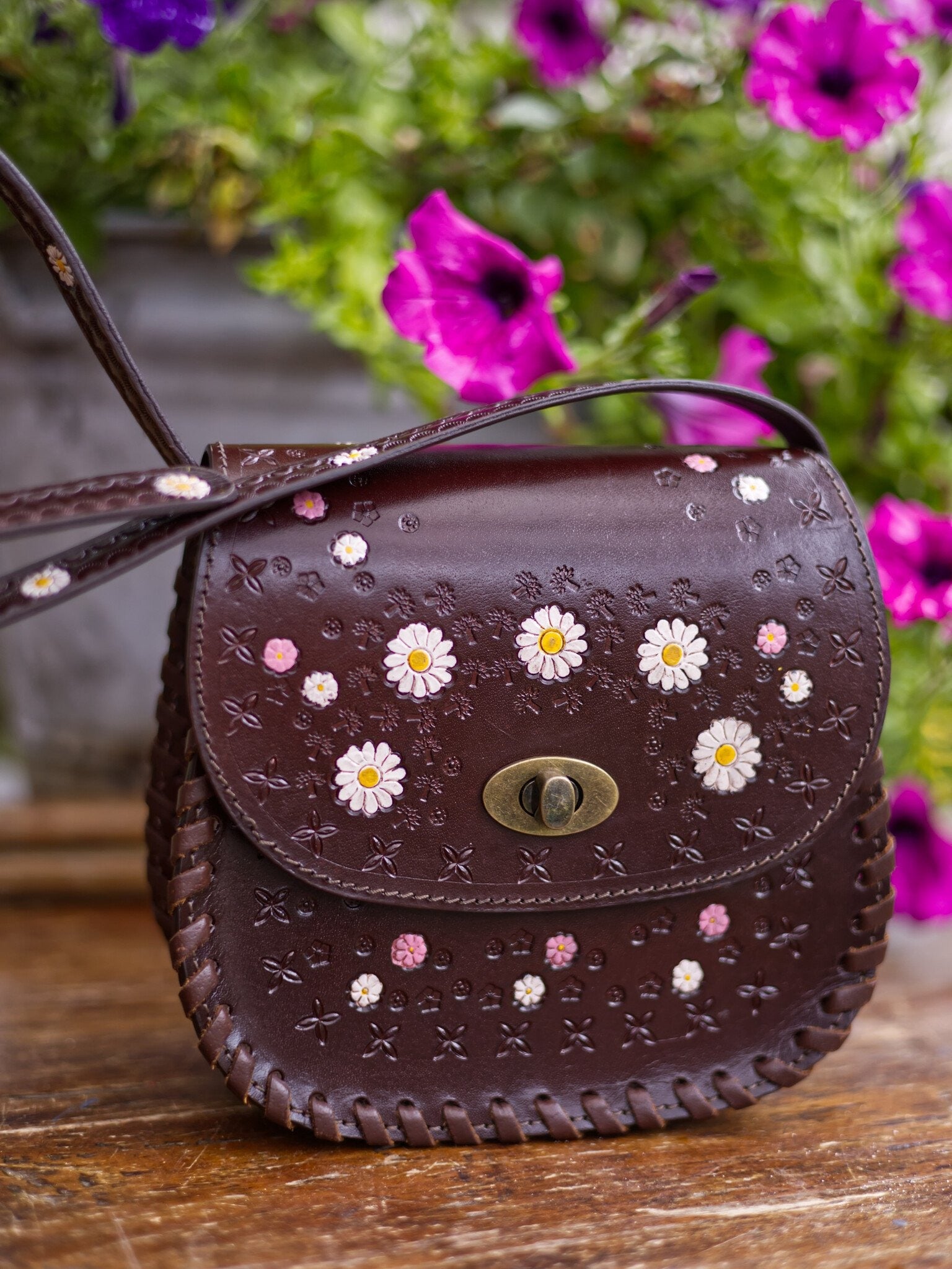 Brown leather handbag with floral patterns on a wooden surface with flowers in the background