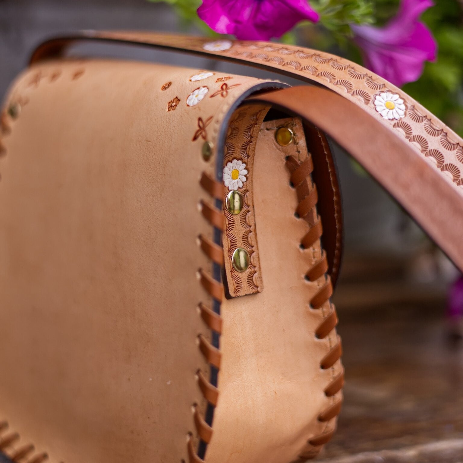 Close-up of a brown leather bag with floral embroidery on a wooden surface with purple flowers in the background.