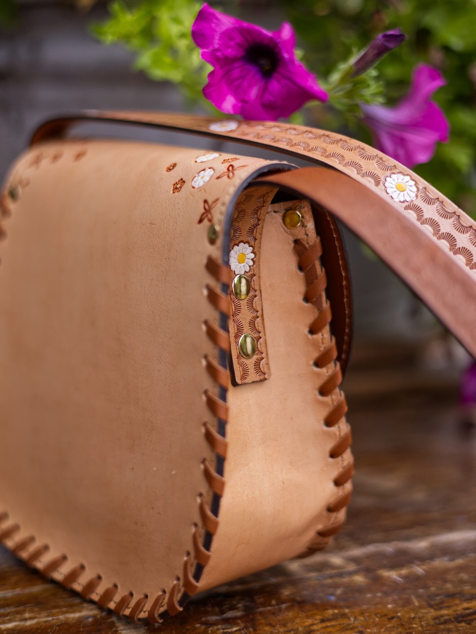 Close-up of a brown leather bag with floral embroidery on a wooden surface with purple flowers in the background.