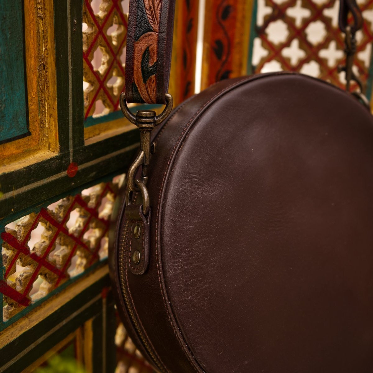 Brown leather round bag with decorative strap against a colorful geometric patterned wall.