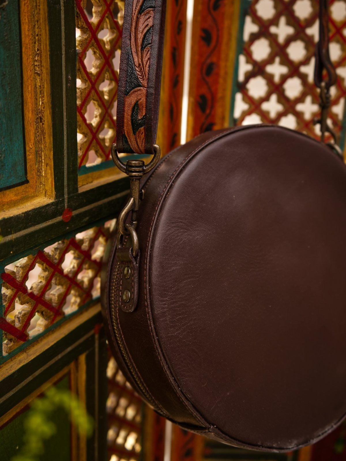 Brown leather round bag with decorative strap against a colorful geometric patterned wall.