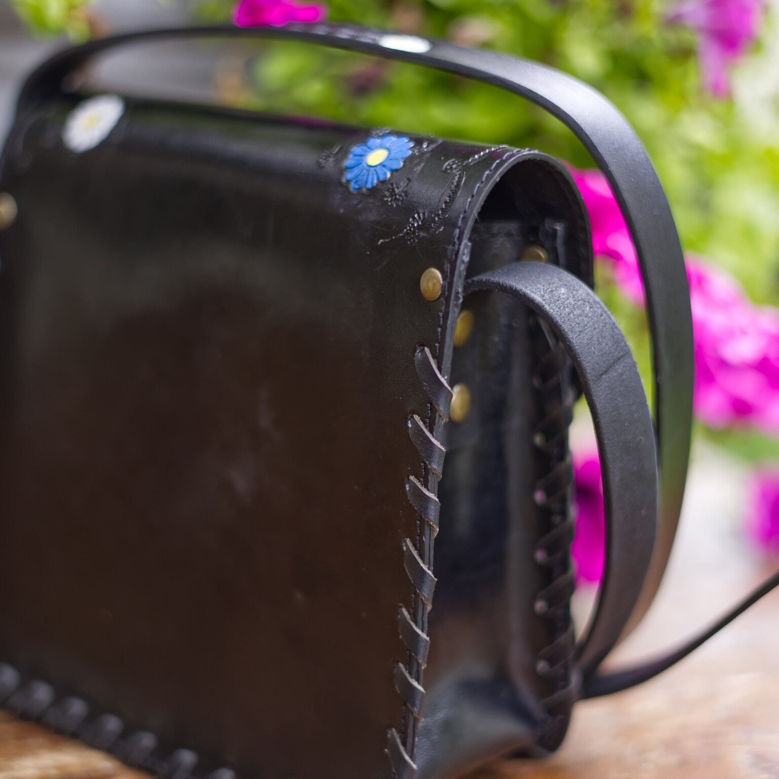 Black leather bag with floral embellishments on a wooden surface with blurred flowers in the background