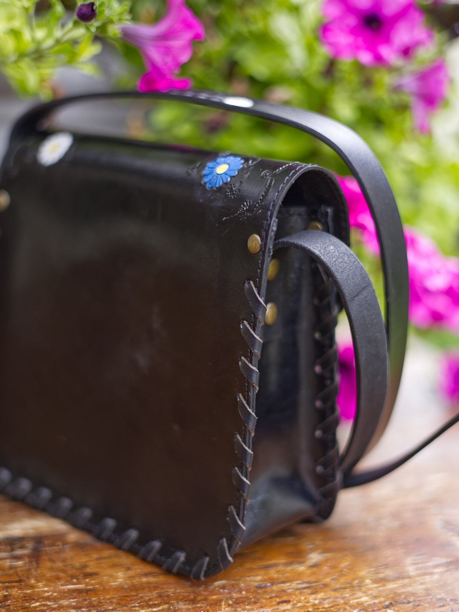 Black leather bag with floral embellishments on a wooden surface with blurred flowers in the background