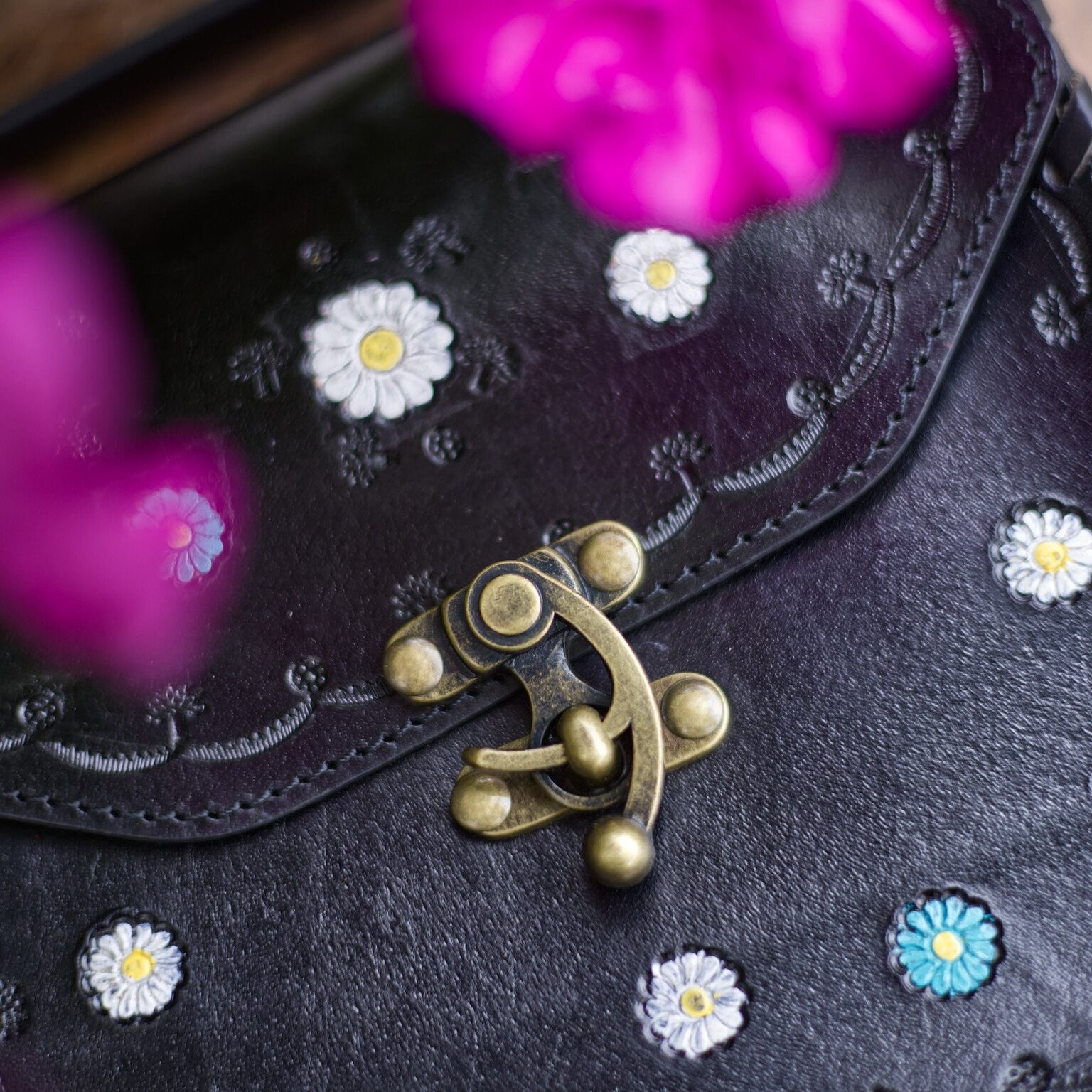 Black handbag with floral embellishments and a gold clasp, with pink flowers in the background.