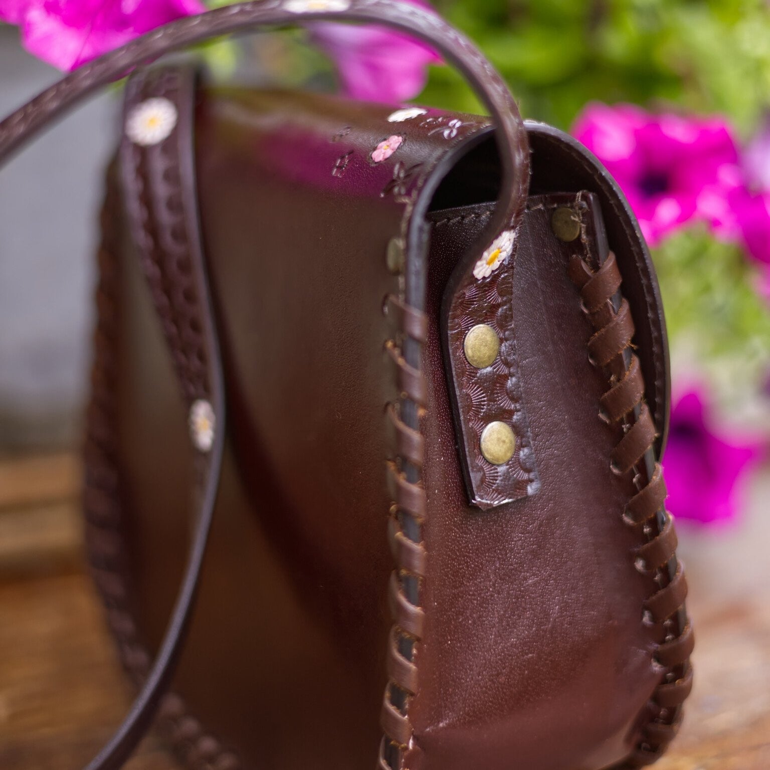 Brown leather handbag with decorative studs on a wooden surface with blurred pink flowers in the background