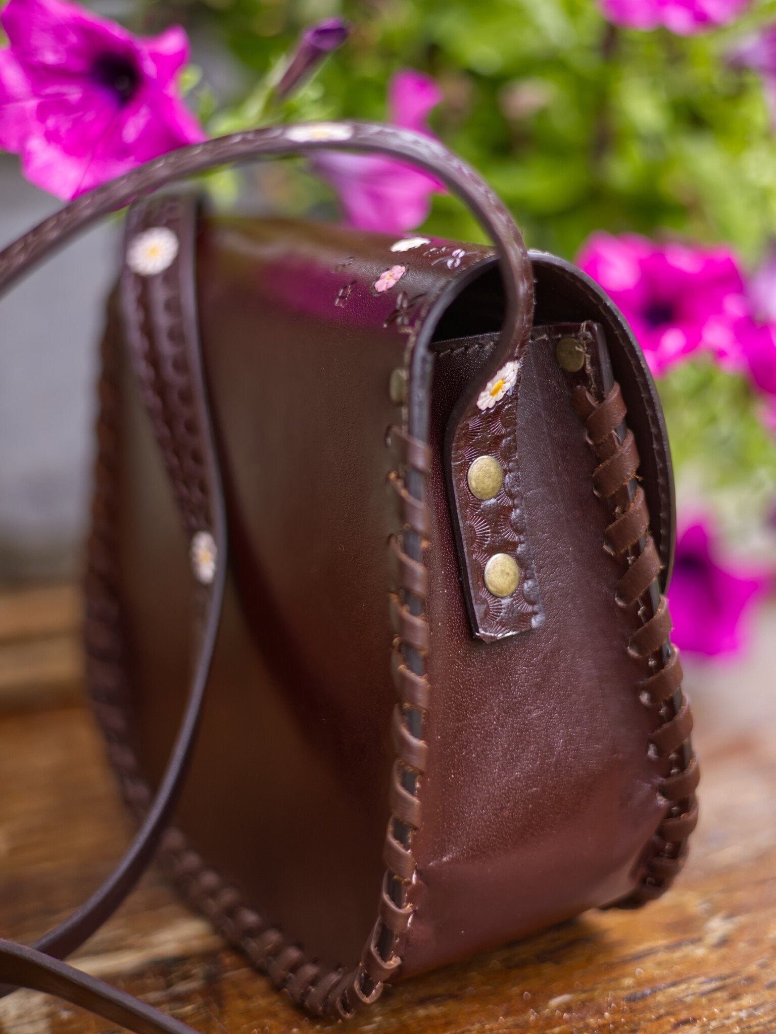 Brown leather handbag with decorative studs on a wooden surface with blurred pink flowers in the background