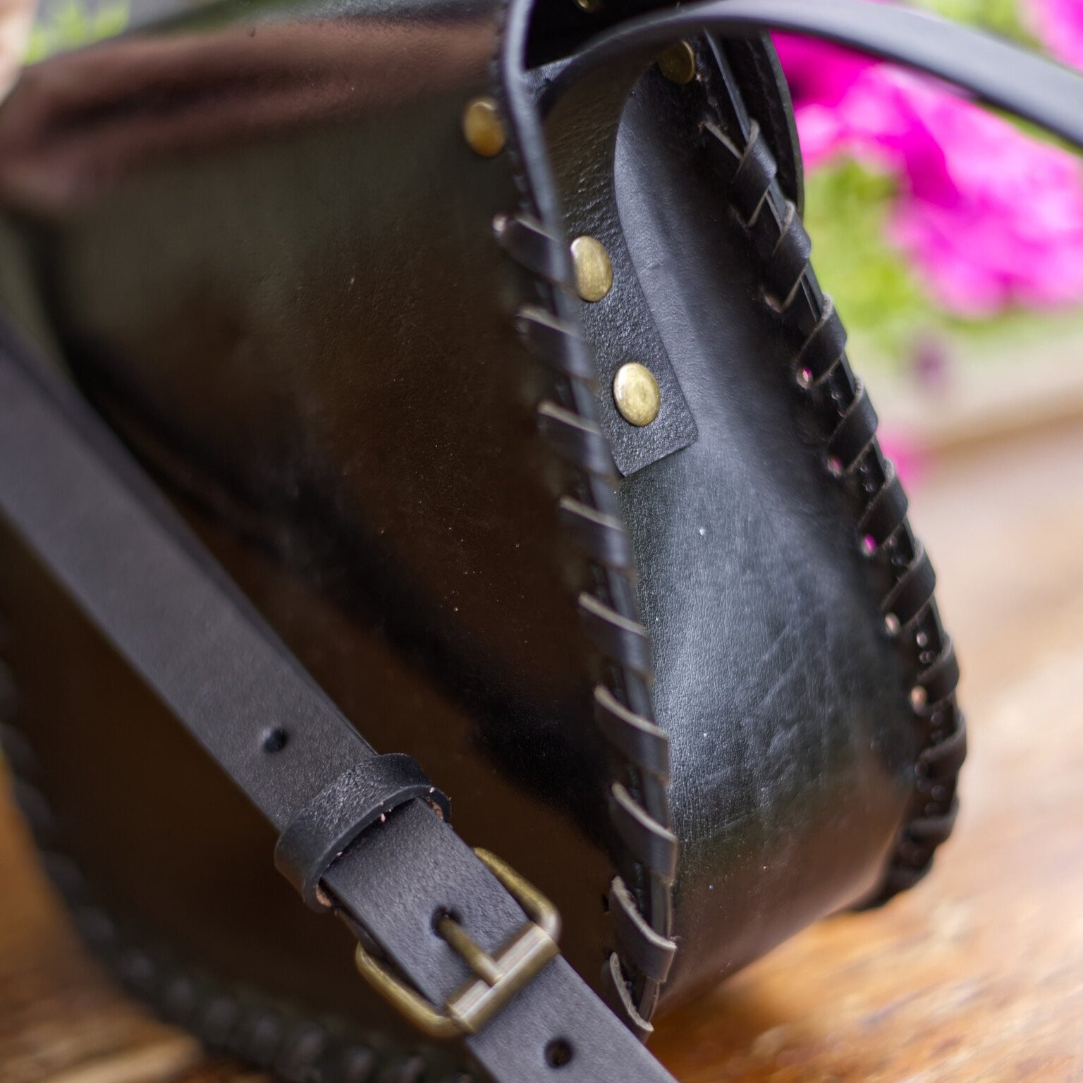 Black leather bag with gold studs and a strap on a wooden surface with blurred flowers in the background
