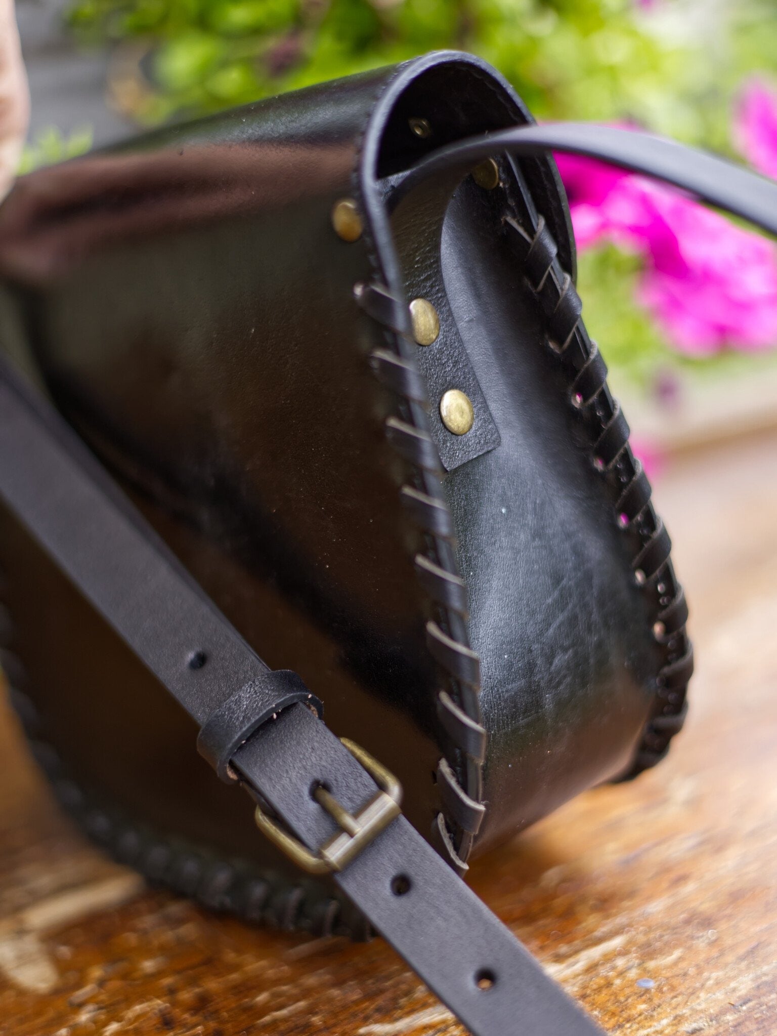 Black leather bag with gold studs and a strap on a wooden surface with blurred flowers in the background