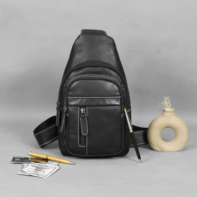 Black leather sling bag on a gray background with a pen, money, and small vase.