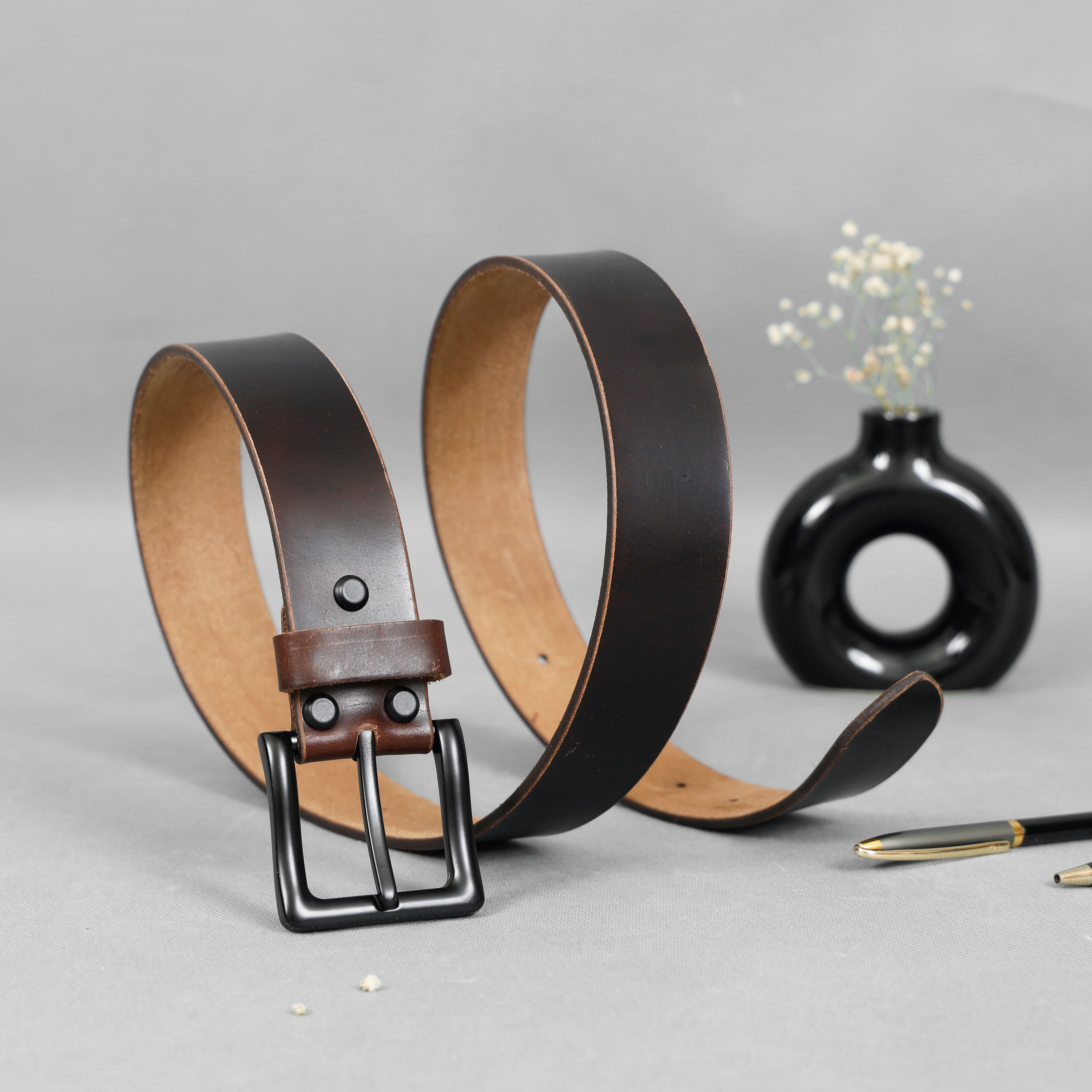 Brown leather belt with black buckle on a gray surface with a black vase and pen in the background.
