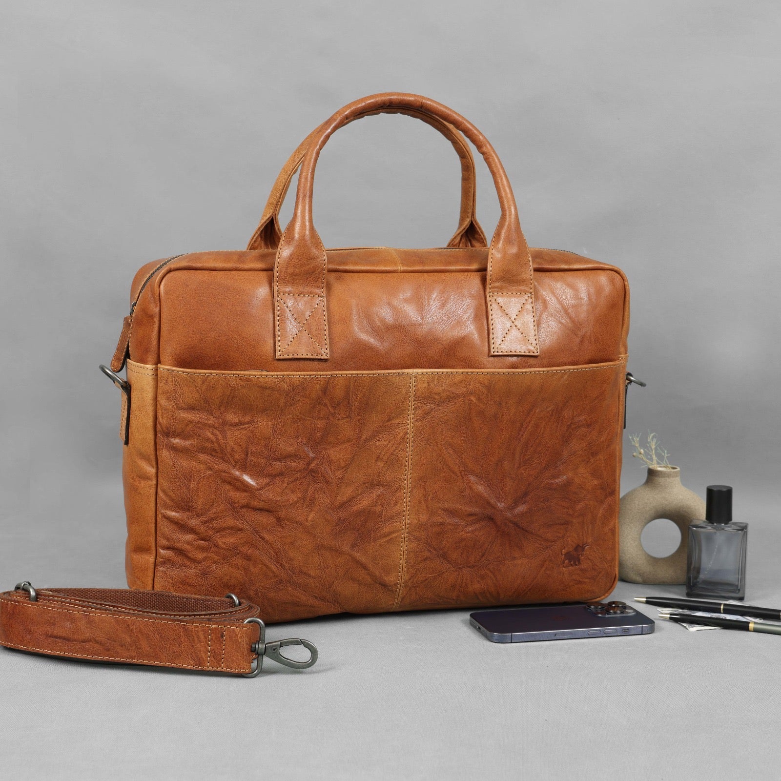 tumbled yellow textured leather laptop bag with a removable shoulder strap, handles, and two front pockets, displayed on a grey surface with a smartphone, pens, and decorative items.