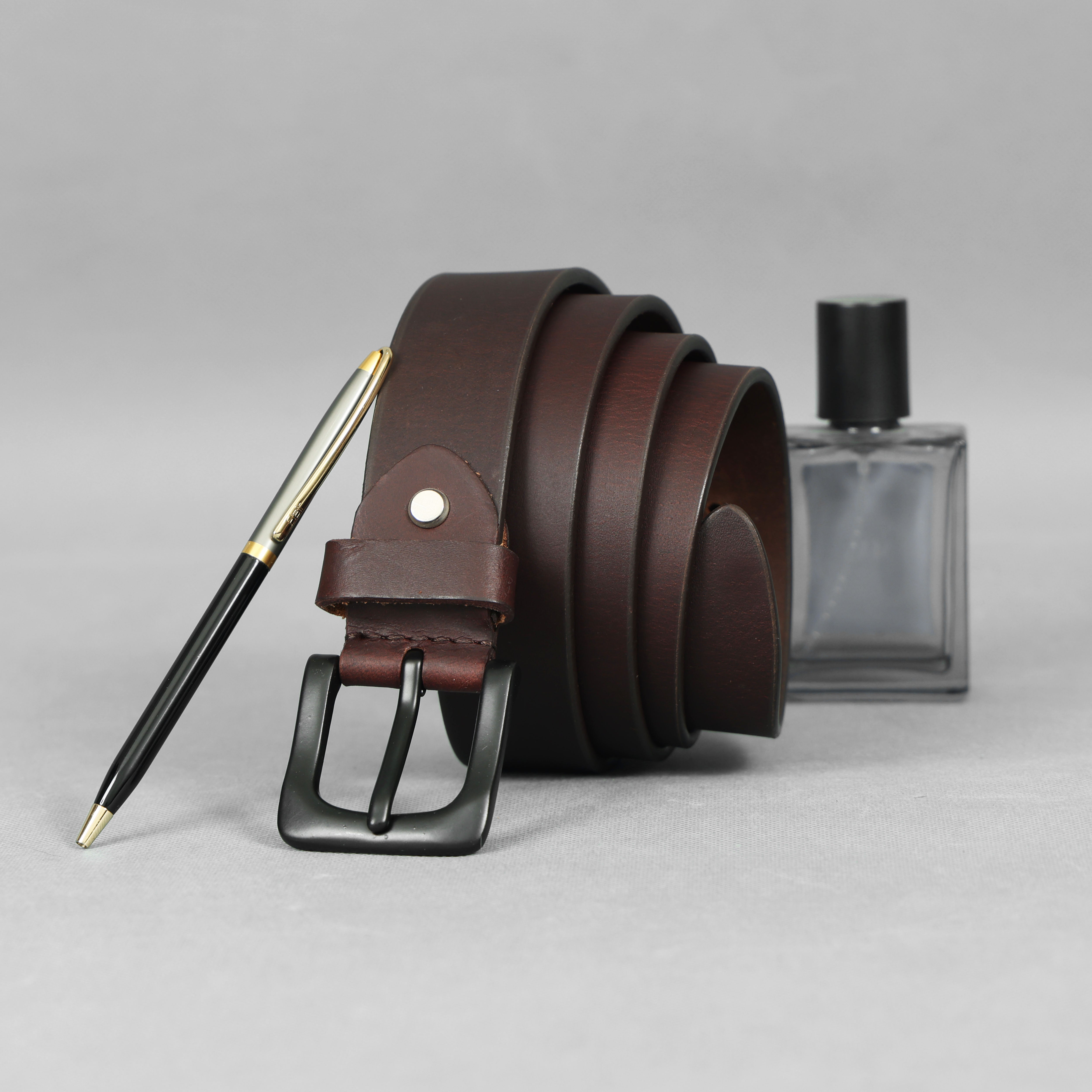 Rolled up Dark Brown Full-Grain Leather Belt for men with a black matte buckle from GM Leather Auckland, displayed next to a gold-tipped pen and cologne
