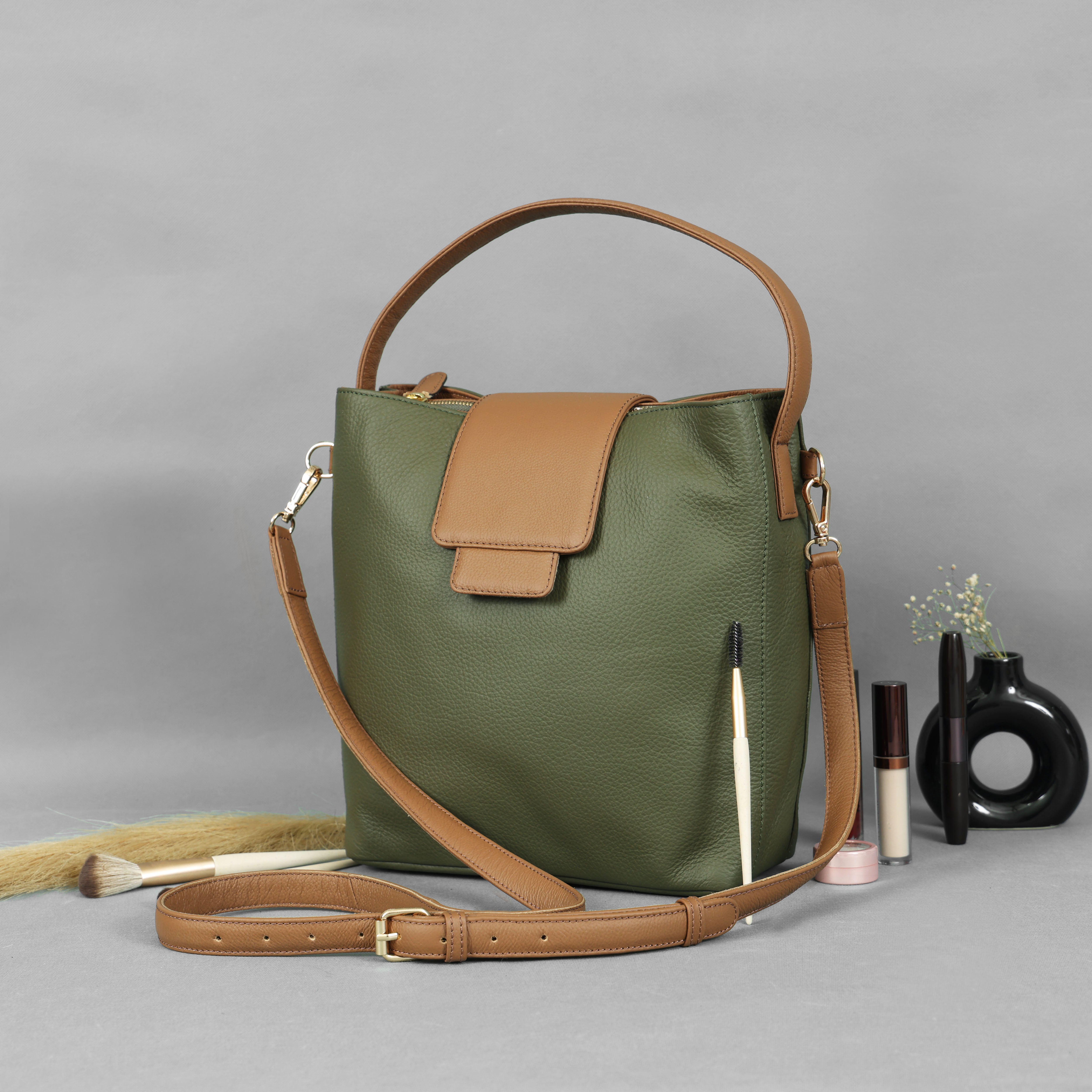 Front view of a two-tone leather bucket bag. The main body is olive green with a tan flap closure, top handle, and adjustable shoulder strap.